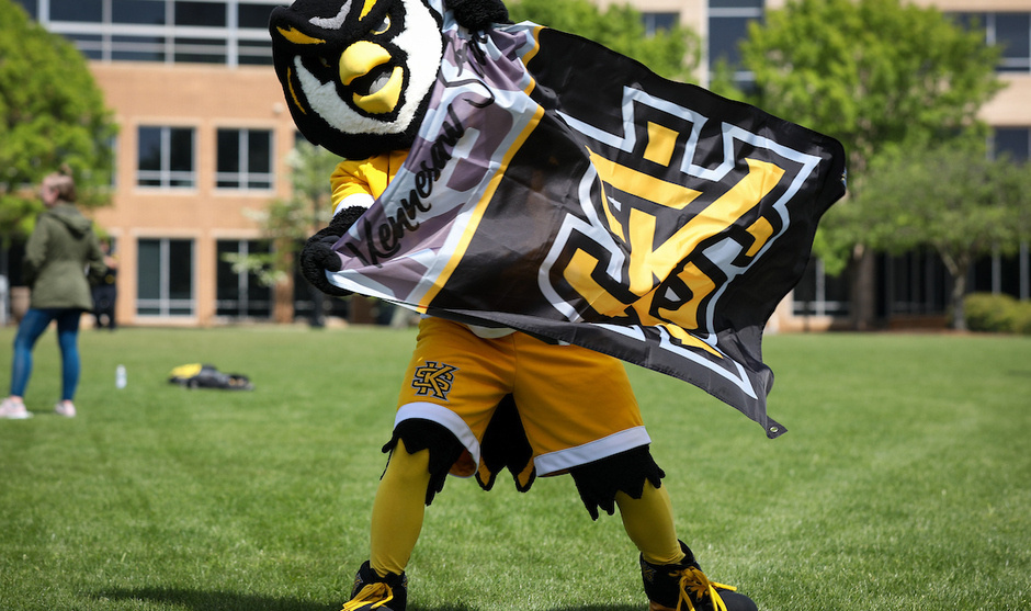 KSU Scrappy mascot wearing black and gold stands on a grassy campus lawn, holding a large KSU flag, with academic buildings visible in the background.