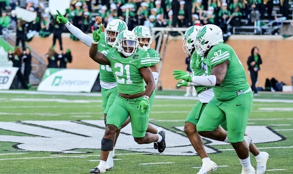 UNT football players on the football field with members of the marching band in the stands.
