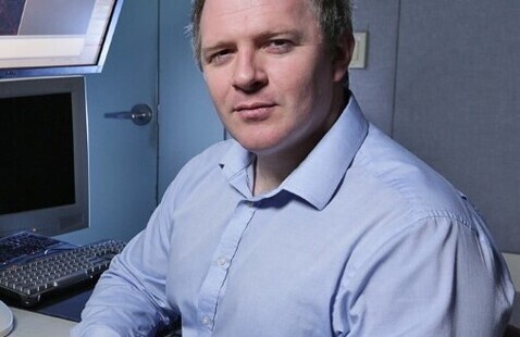 Man in blue shirt smiles slightly, sitting with folded hands and facing the camera