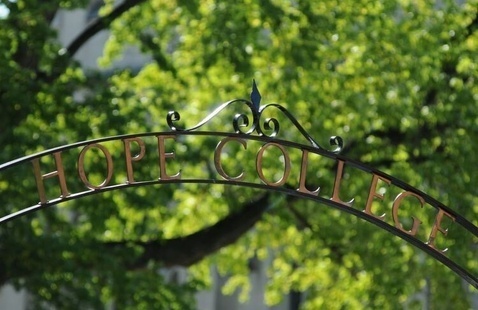 A decorative iron archway with the words "HOPE COLLEGE" in bronze letters, set against a backdrop of lush green trees in full leaf, suggesting a vibrant spring or summer day.