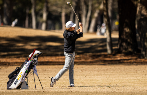 An App State golfer finishes his swing. An App State golf bag can be seen sitting to the left of the golfer.