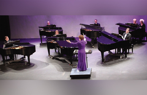 An image of a conductor leading the Six Piano Ensemble performers on stage.