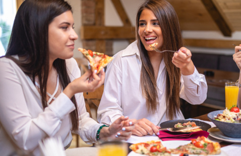 Two students eating pizza in a dining hall, while talking and laughing