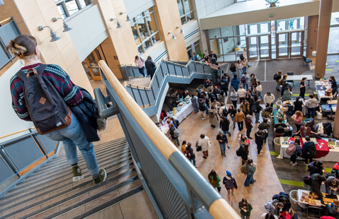 Student walking down a stair case in the Davis Center at the bottom of the stairs there is an event going on with lots of students gathered in the Atrium