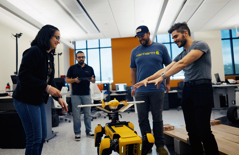 Four people in a lab stand around a legged robot carrying a small drone.