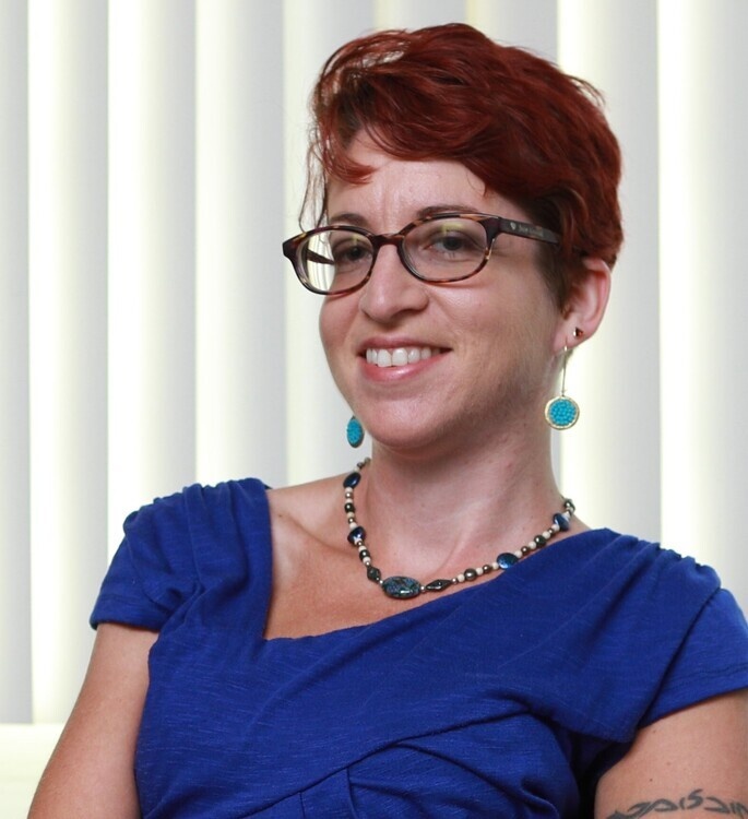 The photo displays a woman with short red hair wearing blue earrings and a blue top. She is wearing glasses and smiling at the camera. 