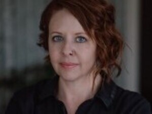 The picture displays a white woman with red hair and a nose ring gazing at the camera. She has a neutral expression and is wearing a black collared shirt. 