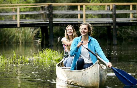 Two students kayaking in a kayak in a body of water with a boardwalk in the background.