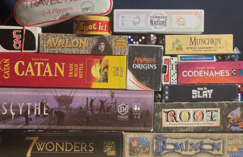 A wall of board games displaying games such as 7 wonders, Dominion, Scythe, Root, Catan, Magic the Gathering, Here to Slay, Codenames, Avalon, Uno, Spot It!, Travel Rummy, Command of Nature, Munchkin, standard decks of cards, and a variety of dice. 