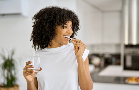 Happy healthy young adult African American woman model talking pill holding glass of water standing in the kitchen at home.
