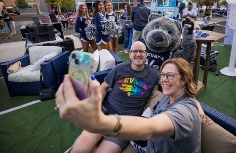 Two individuals taking a selfie photo outside in Midtown with the University of Nevada, Reno mascot. Cheerleaders in the background. 