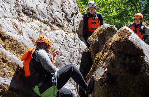 A student in a personal floatation device rappels down a cliff while two other students watch.
