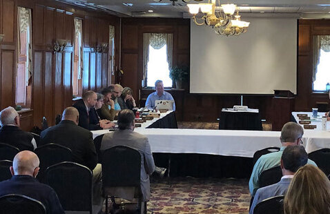 People sitting behind a table in the background with an audience of people sitting in chairs in the foreground