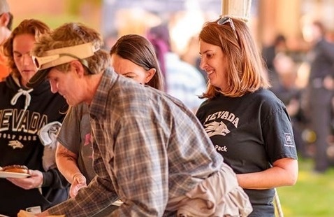 Attendees gather under an outdoor tent, helping themselves to food from buffet trays. Several people wear "Nevada Wolf Pack" apparel, indicating school spirit and community involvement.