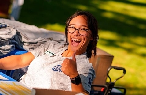 A cheerful individual wearing glasses and a gray shirt with a name tag sits at an outdoor check-in table, smiling and giving a thumbs-up. Behind them are neatly stacked folded shirts, suggesting event merchandise or giveaways.