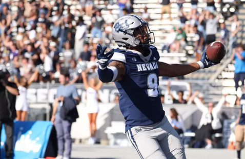 A Nevada football player wearing a navy blue and white uniform with the number 9 celebrates in a stadium, holding a football in one hand and extending both arms outward. The helmet features the word 'Pack.' Behind the player, cheerleaders, teammates, and a crowd of fans in the stands cheer enthusiastically.