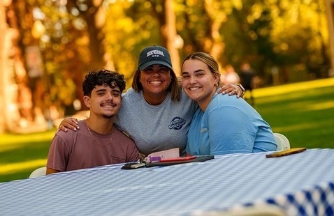 Three individuals sit closely together at a picnic table covered with a blue-and-white checkered tablecloth. They smile warmly at the camera with arms around each other, enjoying a sunny day outdoors among trees and grass.