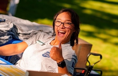 A cheerful individual wearing glasses and a gray shirt with a name tag sits at an outdoor check-in table, smiling and giving a thumbs-up. Behind them are neatly stacked folded shirts, suggesting event merchandise or giveaways.
