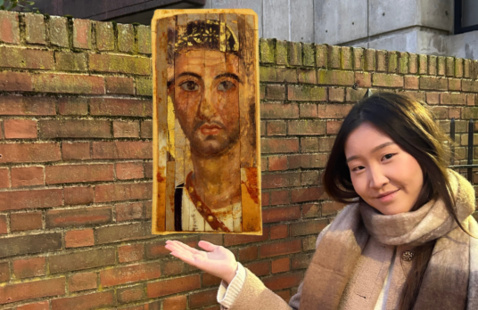 In this photomontage, a young woman, wearing a brown scarf and a beige winter jacket,  gestures toward a portrait that appears to float beside her. The portrait shows a lightly bearded young man and is painted on several slightly mismatched strips of material. 