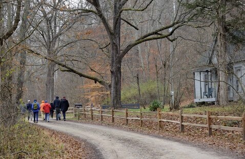 A group of people walk down a gravel road in early November. A wooden fence parallels the road, with a weathered house sitting on the other side.