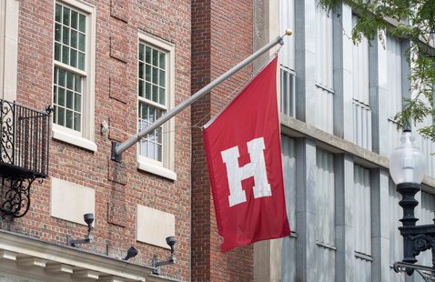 A Harvard flag featuring a bold 'H' in the center hangs beside a brick building.