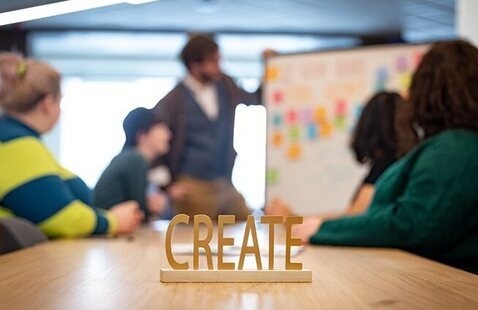 Sign that says "Create" on a table with people around it engaging with a white board with sticky notes.
