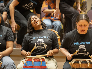 Three MHC students drumming and singing at a performance, dancers in the background.