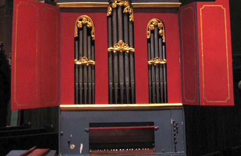 Decorative pipe organ with red and gold panels and ornate gold detailing around the pipes, set inside a church with stained glass windows in the background. The organ has three vertical sections of pipes and two open side doors revealing the pipes.