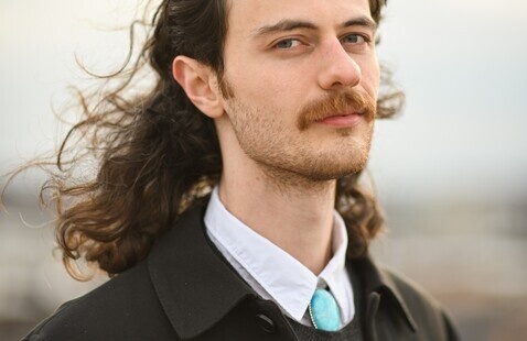Person with long, wavy hair wearing a black coat over a dark sweater and white collared shirt, accented with a turquoise tie. The background is softly blurred, suggesting an outdoor setting with a cloudy sky.