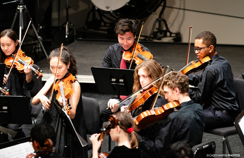 A group of musicians seated on stage performing in a chamber orchestra. Several players are holding violins and violas, reading sheet music on black stands. The musicians are dressed in formal black attire, and the background includes percussion instruments and stage equipment. The photo is credited to Casey Martin Photos.