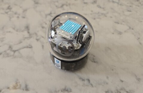 A lit-up Shero BOLT robot sitting in a stand on a white marble background.