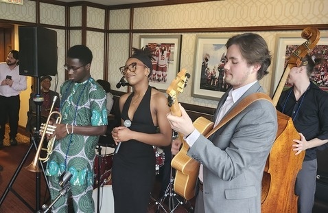 A live jazz band performing in an indoor lounge setting. The group includes a trombone player in a patterned green outfit, a vocalist in a black dress, a guitarist in a gray suit, and a double bass player in dark clothing.