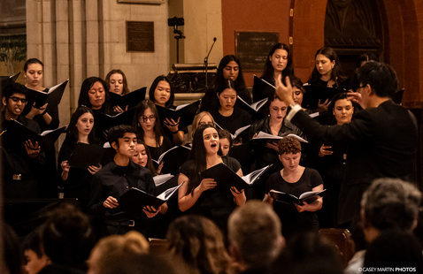 A choir performing inside a historic church or chapel, with singers holding open sheet music and a conductor leading the group. The background features ornate architecture, stone walls, and warm lighting, with an audience seated in the foreground.