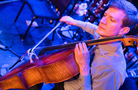Stefan Freund of Alarm Will Sound plays the cello passionately on stage, surrounded by blue lighting. His focused expression and formal attire suggest a live performance setting.
