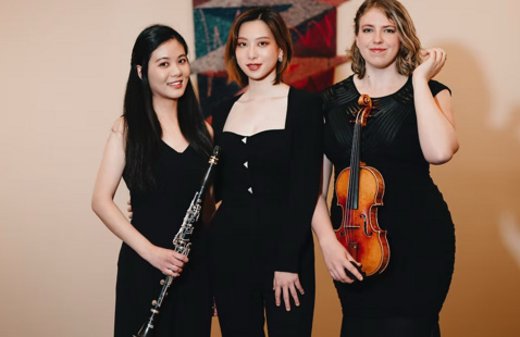 Three women in black dresses pose with instruments, a clarinet and a violin, against an abstract tapestry backdrop, exuding confidence and elegance.