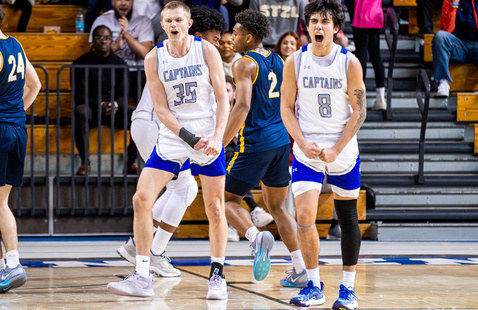 Two male basketball players in white Captains jerseys celebrate on the court, with a player from the opposing team nearby.