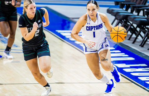 A female basketball player in a white Captains jersey dribbles the ball down the court while being closely guarded by an opposing player in a black jersey.