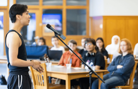 a person standing in front of a microphone performing. There are people seated at wooden tables watching the performance.