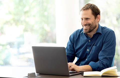Man sitting at a table with large windows behind them and a laptop open in front of them. 