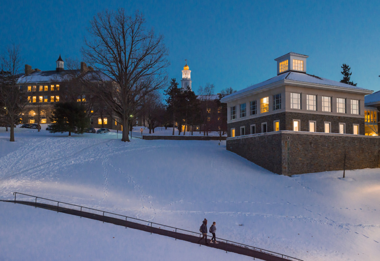 A couple walk a pathway at dusk in front of a snow-covered Colgate campus hill.