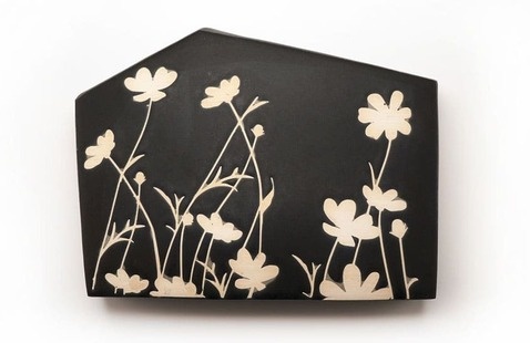 A black ceramic piece with a matte finish, shaped like a house silhouette. The surface features an elegant design of cream-colored wildflowers with long stems and delicate petals, creating a striking contrast against the dark background.