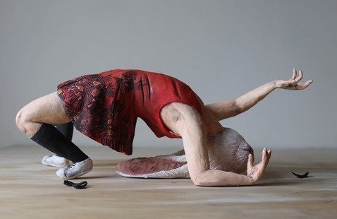 A sculptural figure posed in an arched backbend with arms extended upward. The figure wears a red patterned dress and black knee-high socks, with white shoes. The head is replaced by an elongated, organic form resembling a shell, creating a surreal effect.