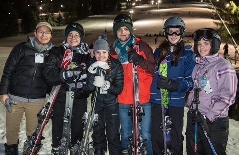 A group of students pose together with their skiing and snowboarding equipment on a night trip to a mountain.
