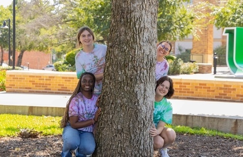 A group of students hanging out on campus, hiding behind a tree.