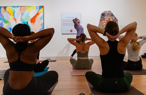 Five women are seated in the gallery practicing yoga. Two artworks can be seen on both sides of the instructor.