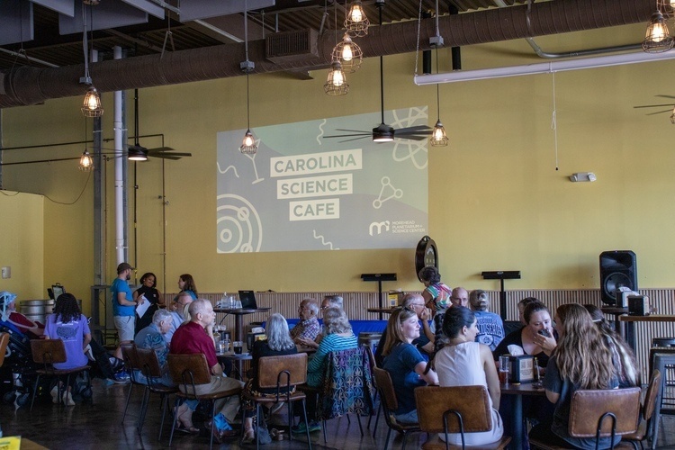 A group of people sit at Haw River Tap & Table. The Carolina Science Cafe artwork is projected on the wall.