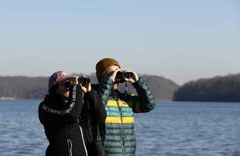 A woman and a man stand with binoculars held up to their faces. A lake is in the background.