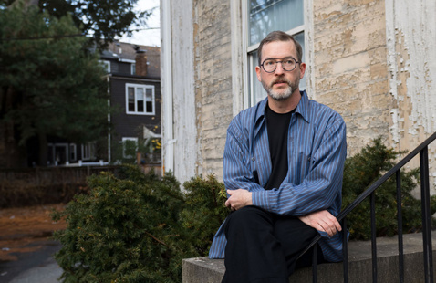 A person wearing glasses, a blue striped shirt, and black pants sits on the steps of an older building with peeling paint, arms crossed and legs crossed, looking slightly off to the side as trees and neighboring houses appear in the background.