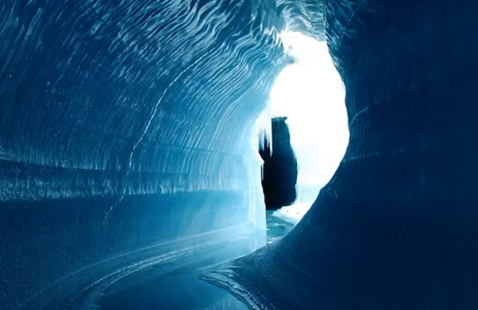 Inside an icy blue glacier tunnel with smooth, rippled walls, looking toward a bright opening where sunlight illuminates the passage.