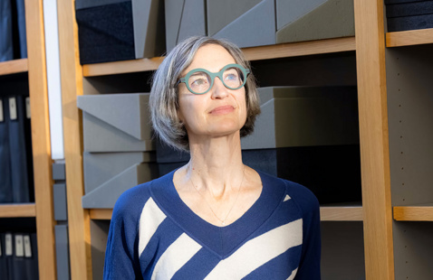 A woman with short gray hair and round green glasses stands indoors in front of shelves filled with archival boxes, wearing a blue sweater with white diagonal stripes and looking slightly upward with a thoughtful expression.